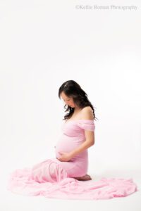 milwaukee maternity photographer. women on ground infront of a white backdrop. she's sitting with her butt on her feet while holding onto her pregnant belly and looking down. she's in front of a white backdrop. she has a long pink dress on that is off the shoulder.