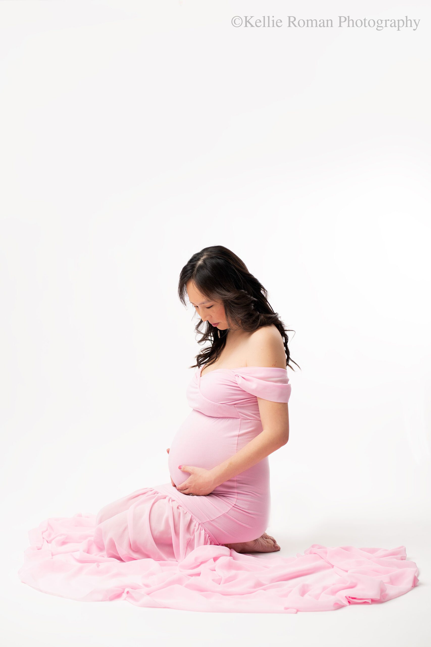 milwaukee maternity photographer. a pregnant women is sitting on her legs on the floor in front of a white backdrop. she has both hands on her pregnant belly and is looking down. she has a long chiffon pink dress on, and has long black hair. 