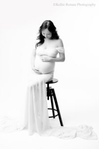 Milwaukee maternity photographer. image is in black and white. women is in long chiffon dress with long train. she is sitting on a black stool in front of a white backdrop. women has long black hair and has both hands on her pregnant belly. she is looking down.