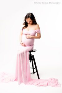 Milwaukee maternity photographer. women is in long pink chiffon dress with long train. she is sitting on a black stool in front of a white backdrop. women has long black hair and has both hands on her pregnant belly. she is looking down.