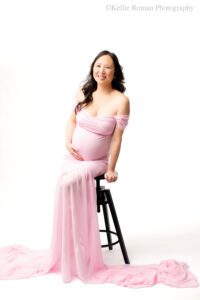 milwaukee maternity photographer. pregnant women is sitting on black stool in front of a white backdrop. women has a very long pink dress on with a chiffon train. the. dress is off the shoulder. women has long dark hair and is holding onto the stool with one hand, and her pregnant belly with the other hand.