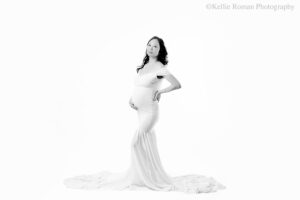 milwaukee maternity photographer. women standing in front of a white backdrop with gown on that is white. the entire image is in black and white. dress has a long train. women has one hand on pregnant belly and the other is on her hip.