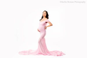 milwaukee maternity photographer. pregnant women is standing in front of a white backdrop. she has a long pink gown on that is off the shoulder. dress has a long chiffon train. women has one hand on her pregnant belly and the other is on her hip. she has long black hair.