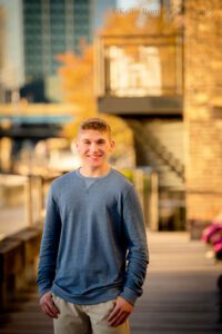 senior session in milwaukee. high school senior boy standing along milwaukee riverwalk on a wood sidewalk. he has khaki pants on with a blue long sleeved shirt. he has his thumbs in his pockets. the buildings behind him are lite up as the sun is setting.