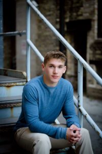 senior session in milwaukee. high school senior boy from greendale is sitting on old rusty metal stairs in Downtown Milwaukee ally. he has khaki pants on with a long sleeved blue shirt. he's leaning forward with his arms resting on his knees and folding his hands.