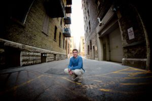 senior session in milwaukee. high school senior boy from greendale is squatting in alleyway in Downtown Milwaukee. he has khaki pants on with a long sleeved blue shirt. there are building surrounded him with old brick.