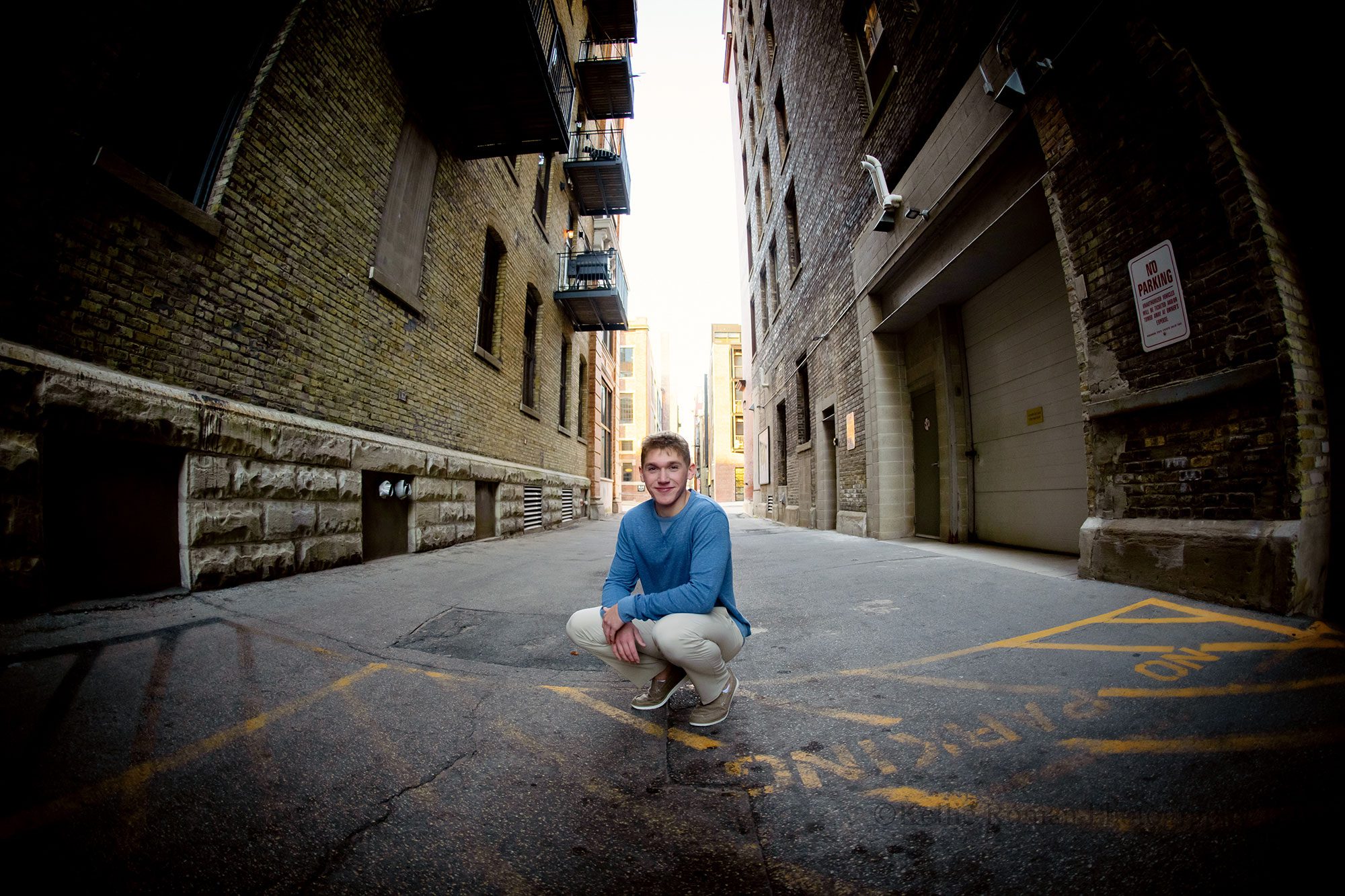 senior session in milwaukee. high school senior boy is squatting in a Downtown Milwaukee ally. he has khaki pants on with a long sleeved blue shirt. 