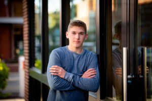 senior session in milwaukee. a high school senior boy is leaning against a glass and metal building in Downtown Milwaukee. his arms are crossed and he has a blue long sleeved shirt on.