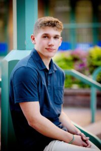 senior session in milwaukee. high school senior boy from greendale is sitting on a concrete in Downtown Milwaukee. he's in front of green and purple flowers, and has khaki pants on and a navy polo shirt. his hands are resting in his lap.