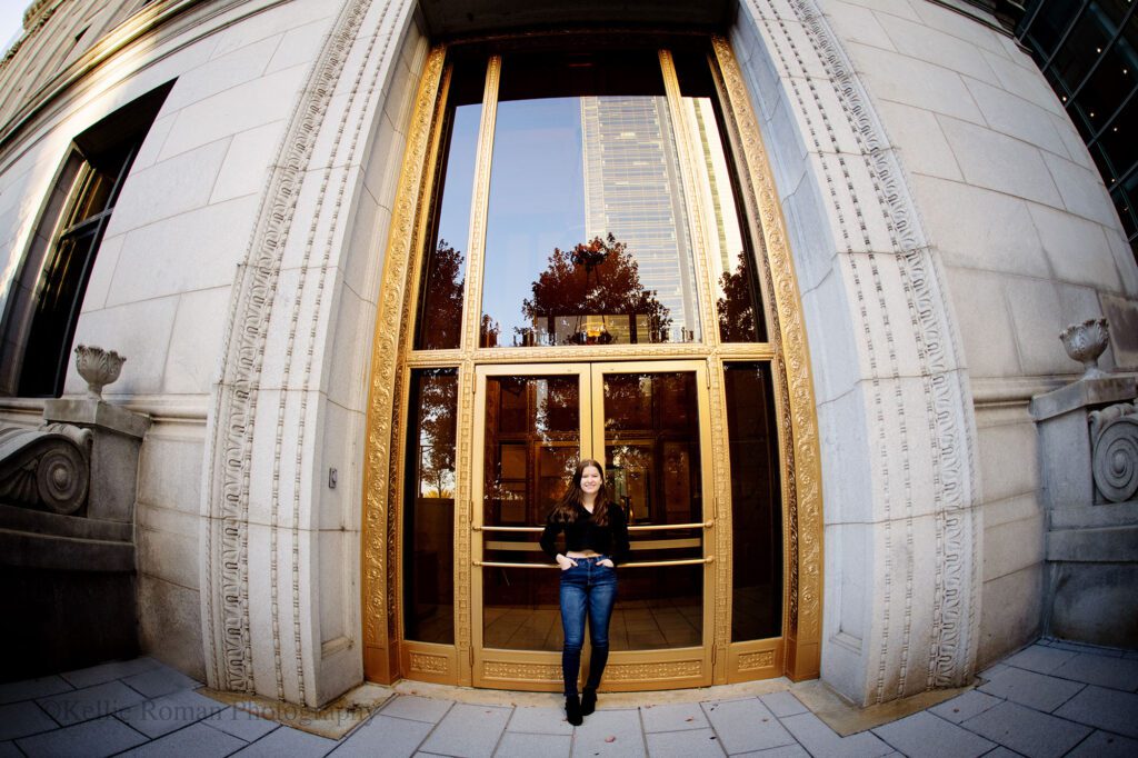 milwaukee senior photographer. kenosha senior girl is standing in front of double gold doors in Downtown Milwaukee. the building has very intricate details. girl is wearing a black shirt and jeans. 

