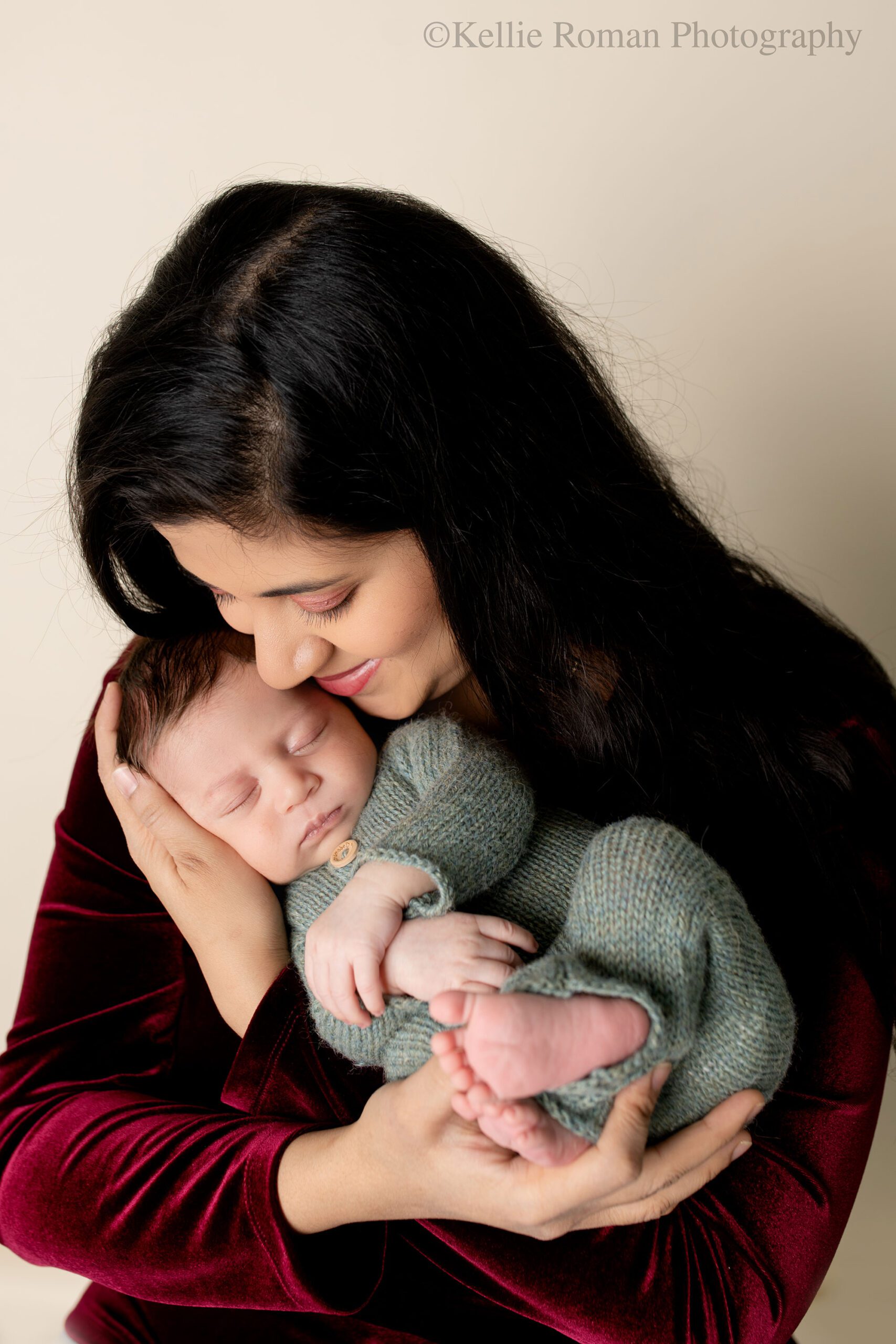 Newborn photographer in milwaukee. a mother is holding her newborn son up near her face and is snuggling with him while closing her eyes. the newborn boy is wearing a green romper with buttons. mom has a marroon velvet shirt on and has long black hair.