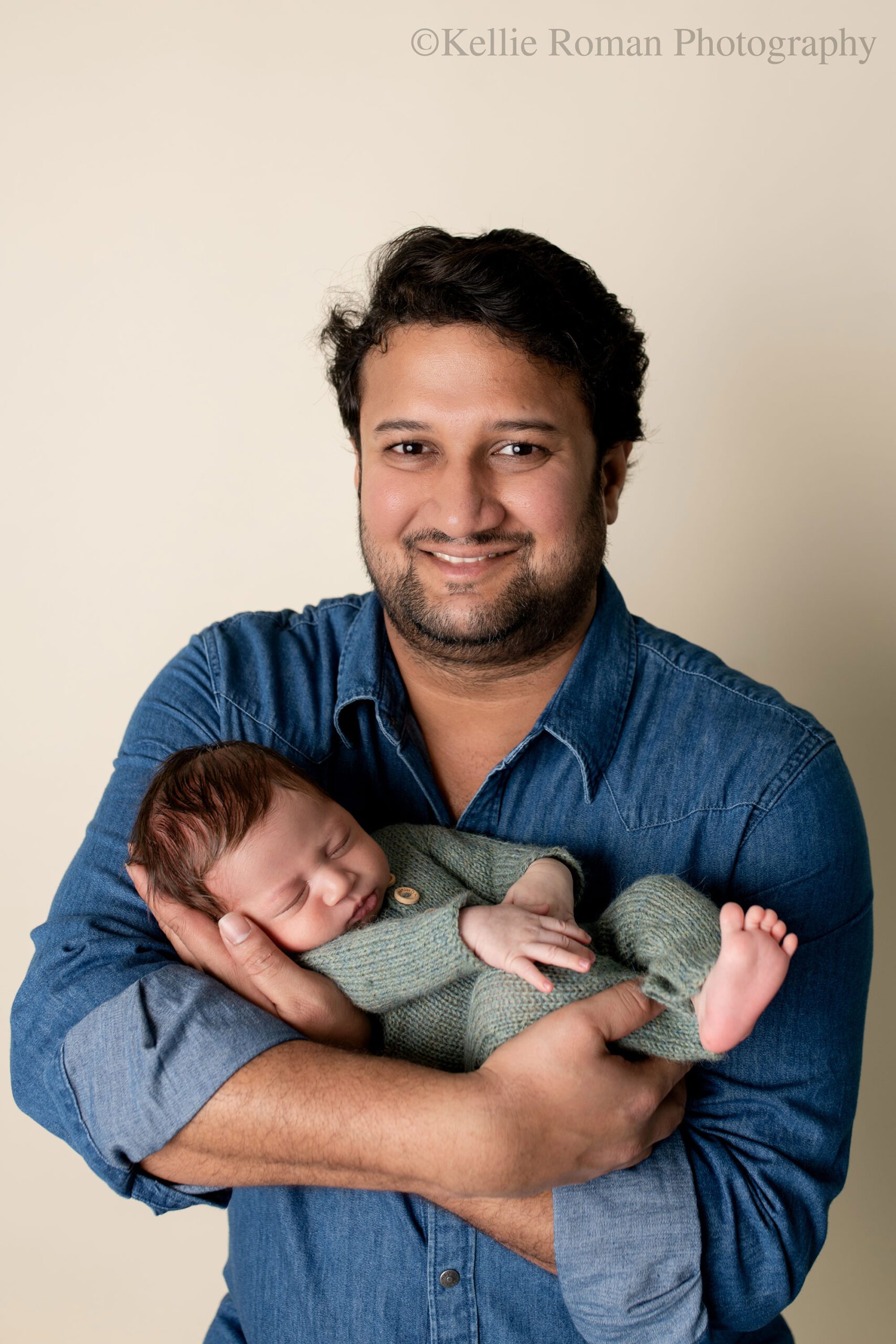 newborn photographer in milwaukee. a father is holding his newborn son and is looking and smiling at the camera. the baby boy is wearing a green knit romper and is sleeping in dads hands. the father has dark hair and a denim shirt on.