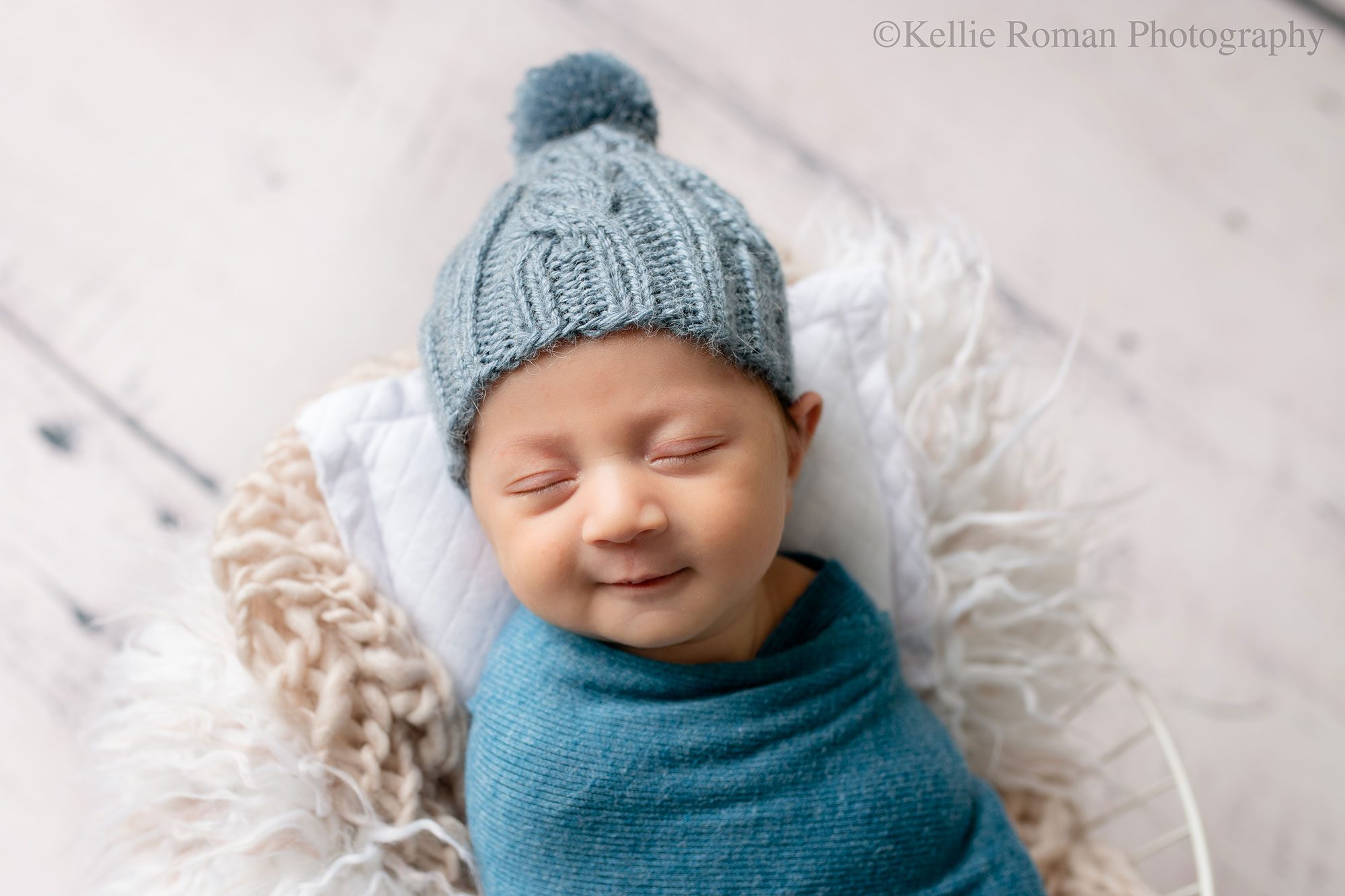 newborn photographer in milwaukee. a newborn boy is swaddled in a blue fabric with a knit pom pom blue hat on. he's on his back sleeping in a basket with a white pillow and beige fluff. the newborn boy is smiling. 