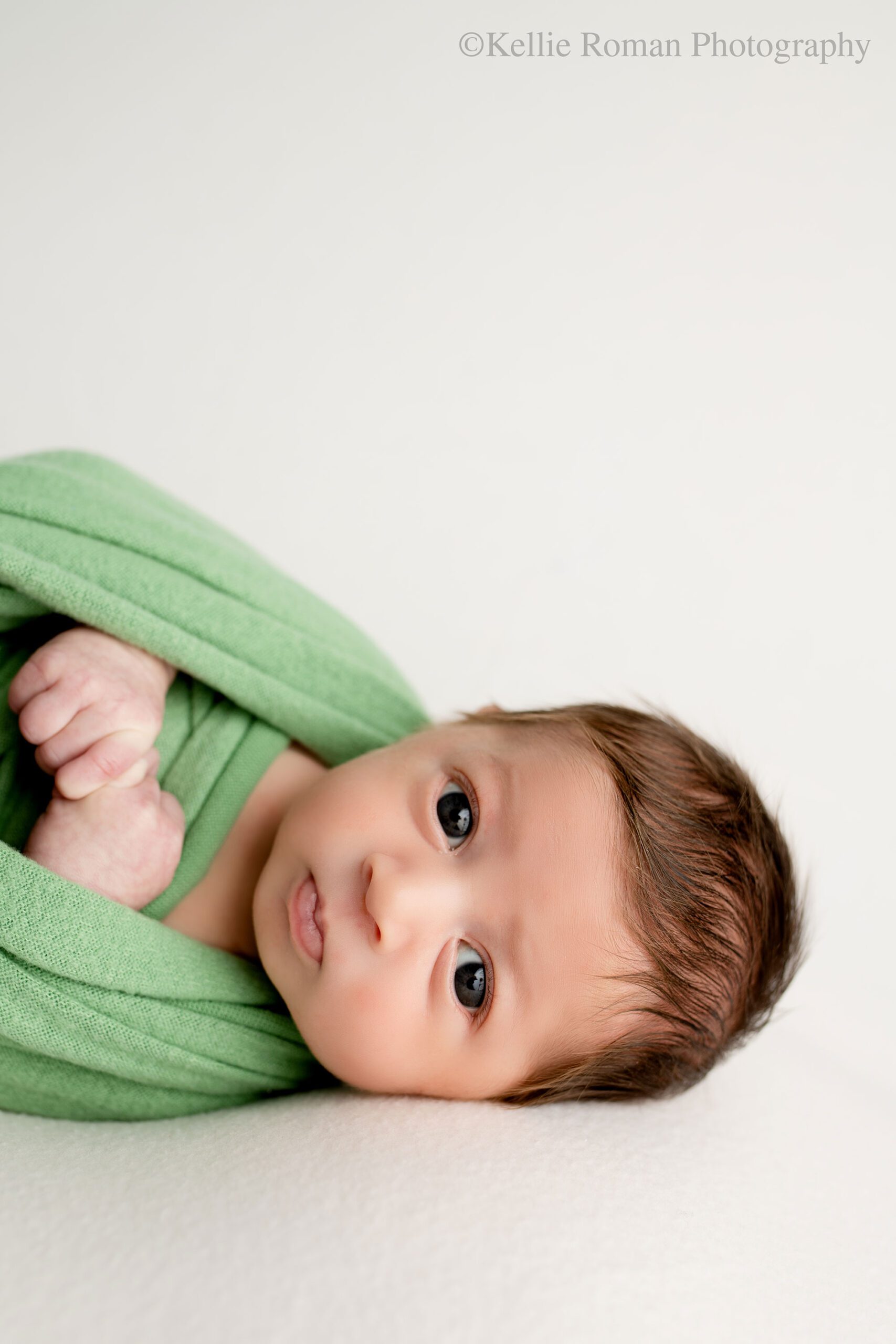 newborn photographer in milwaukee. a newborn boy is wide awake with big round brown eyes. he's looking right into the camera. he has a green swaddled on but his hands are showing. he has dark brown hair.