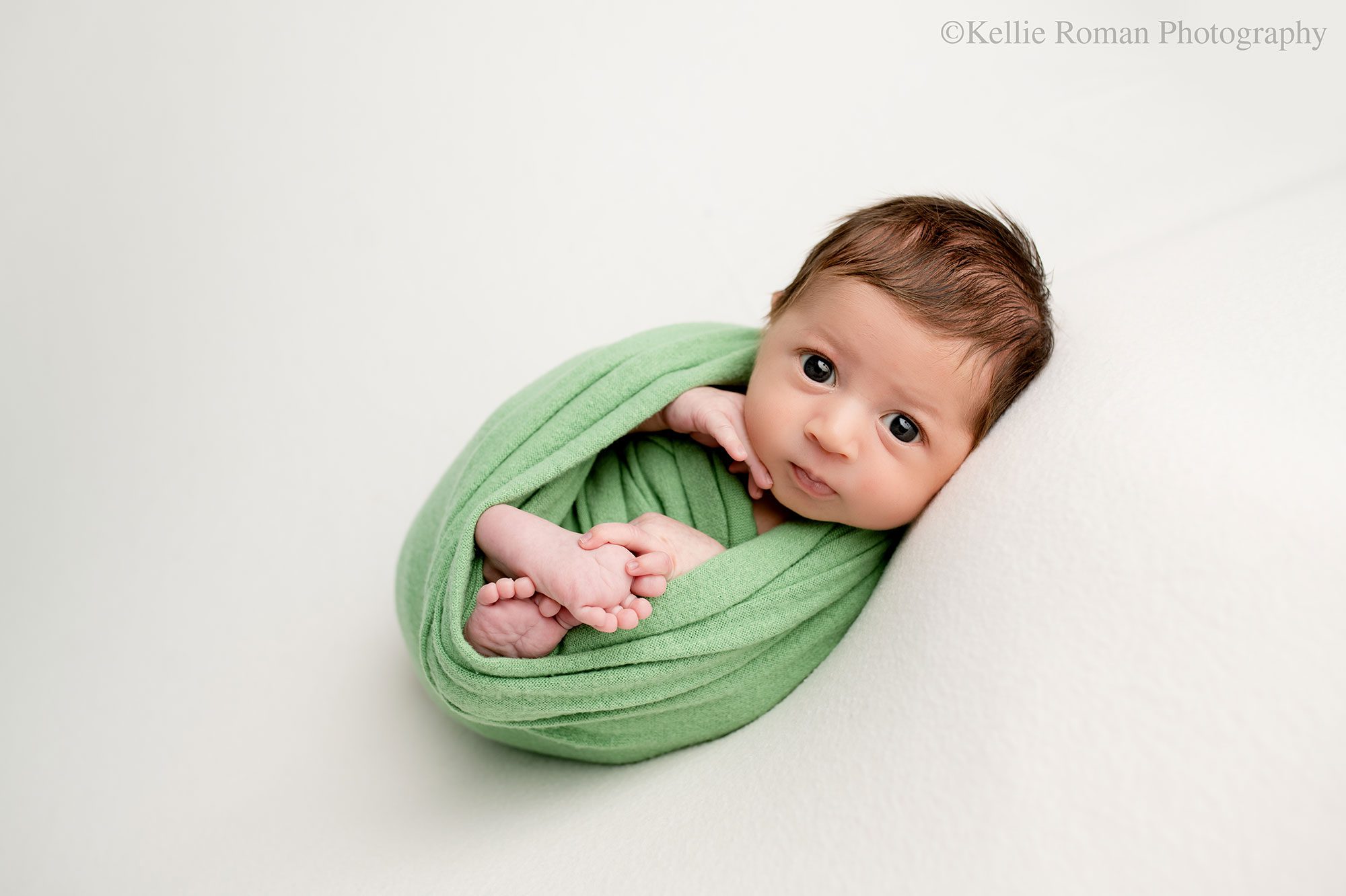 newborn photographer in milwaukee. a newborn boy is wrapped in a green swaddle with his feet and hands sticking out. he has brown hair and big round brown eyes that are open and looking right at the camera. he's on top of a cream colored fabric.