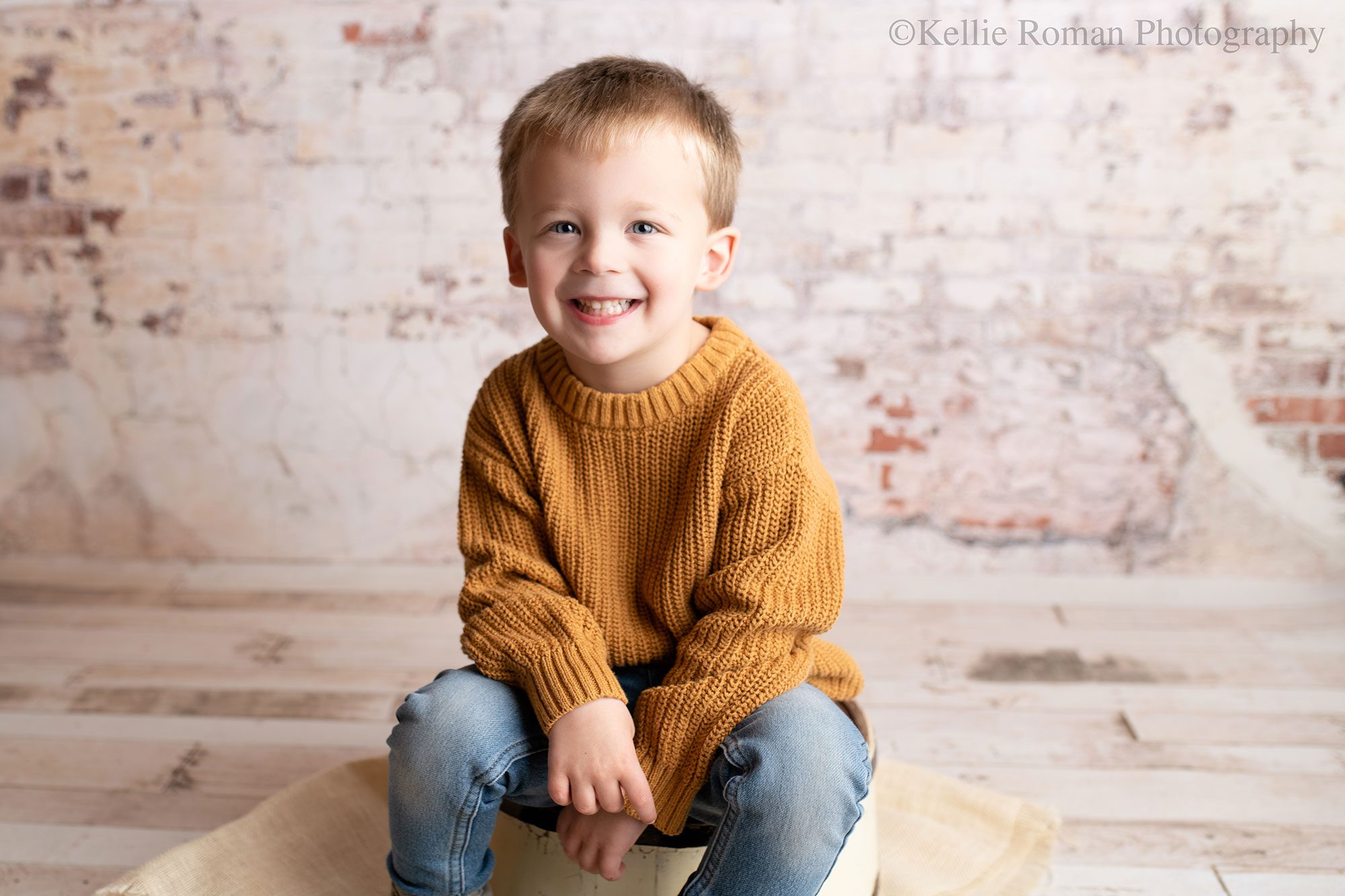 milwaukee child photographer. a boy is sitting on a white tub in front of a cream, brick backdrop. he's smiling at the camera, and has an orange knit sweater on and jeans. he has blonde hair. 