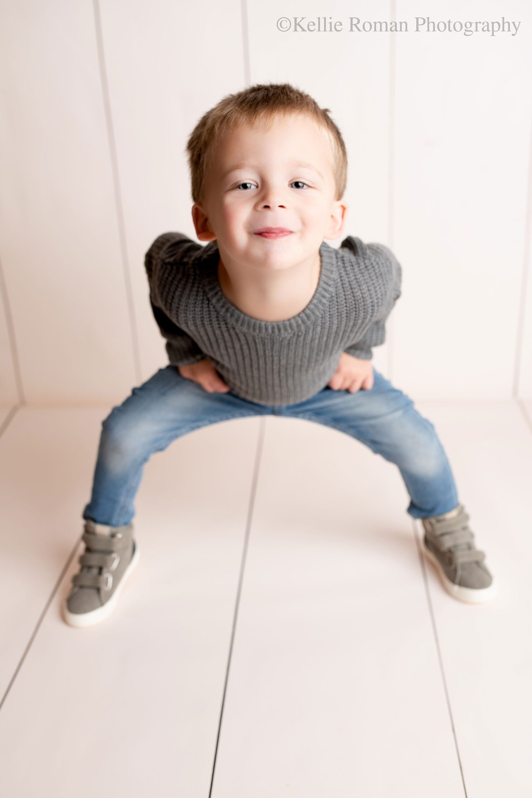 Milwaukee child photographer. a young boy is standing in front of cream striped backdrop with his legs apart and his knees bent. he's smiling at the camera. he has on jeans with a grey knit sweater and grey shoes. 