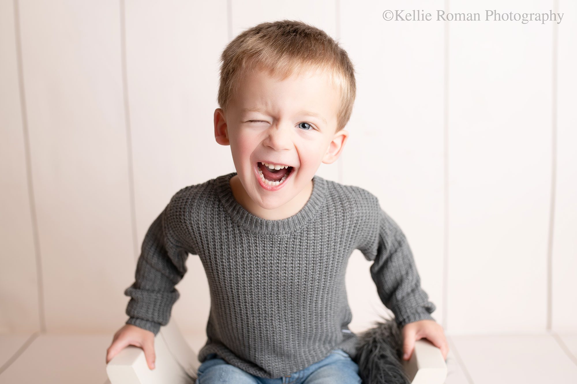 milwaukee child photographer. young boy sitting on a white wood bench and holding onto it. image is a close up of the boy smiling and winking at the camera. he has a grey knit sweater on and has blonde hair. the backdrop is cream, striped.