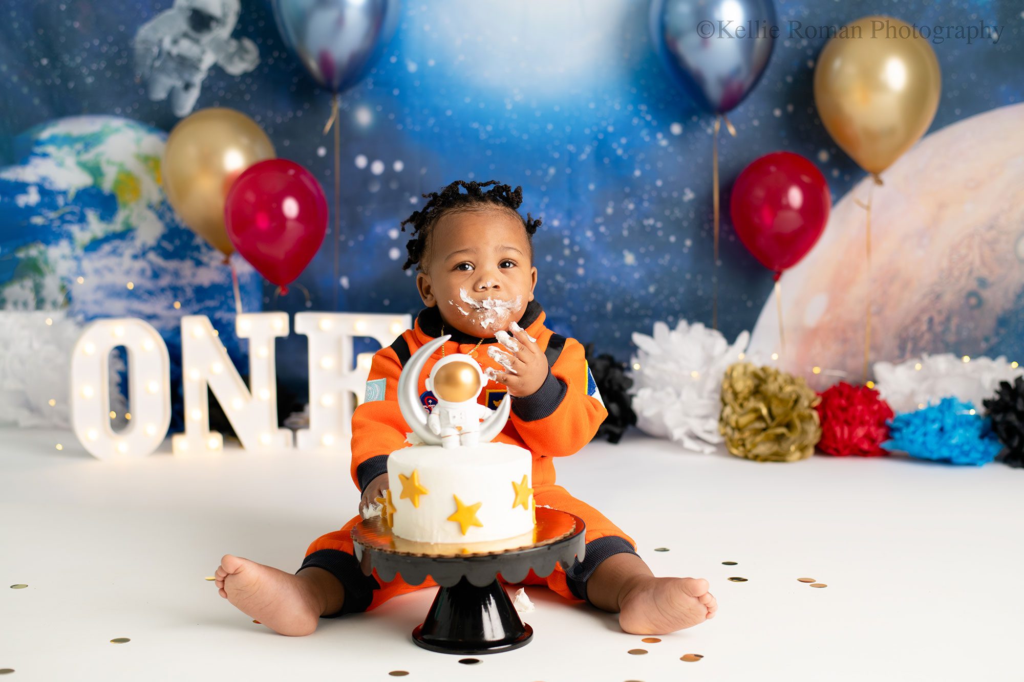 milwaukee cake smash pics. one year old boy is dresses in an orange astronaut onesie sitting behind a white cake with gold stars and a spaceman on a moon cake topper. the backdrop is blues with white stars and blue gold and red balloons. there is a white marquee light that spells out one. the boy has frosting on his face and on his hands.