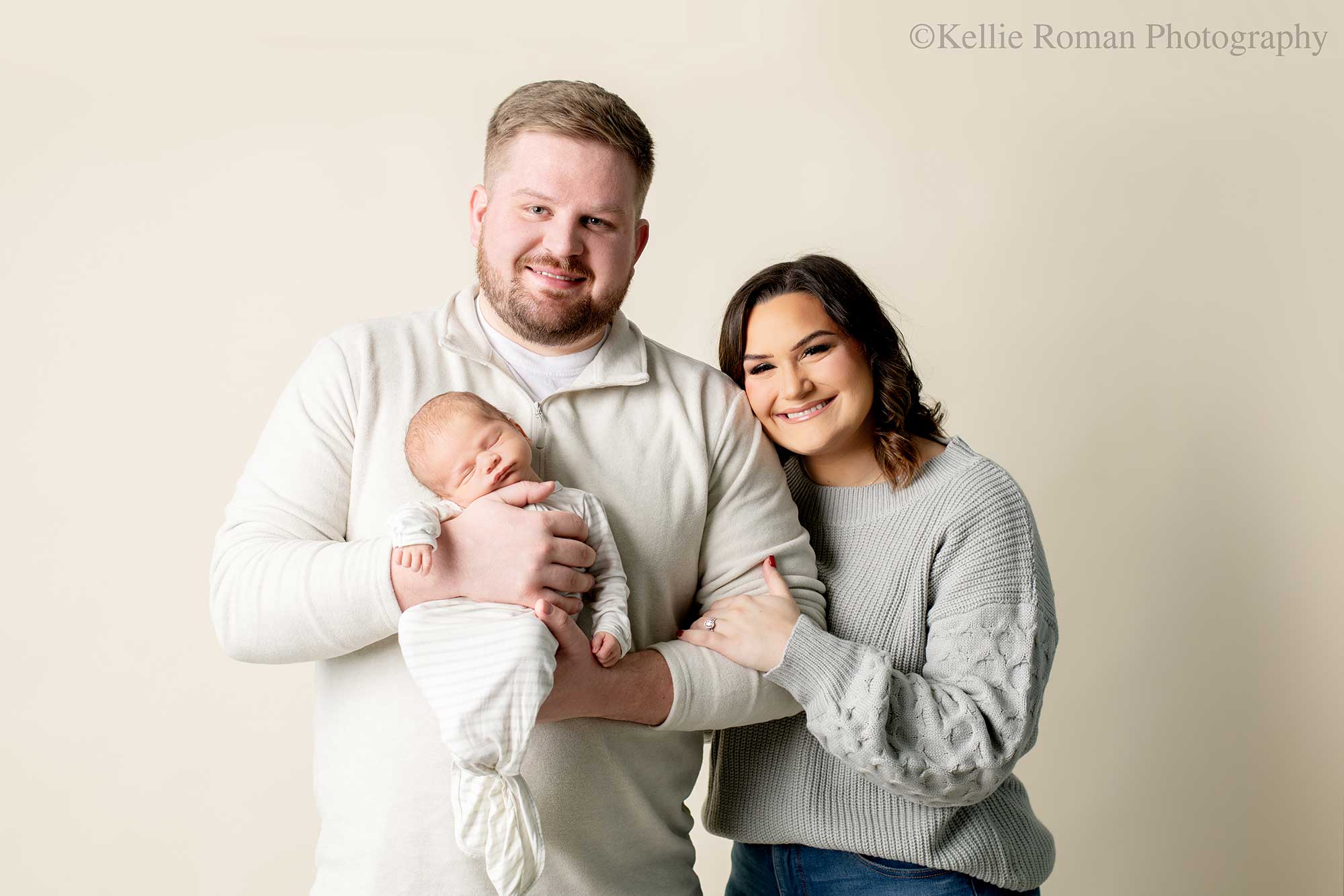 milwaukee newborn photographers. a husband and wife are standing in front of cream backdrop smiling and looking at the camera. the husband is holding their newborn baby against his chest, while he is sleeping. the mom is leaning against dads arm with her hand on his arm. the family is wearing shades of cream and light grey.