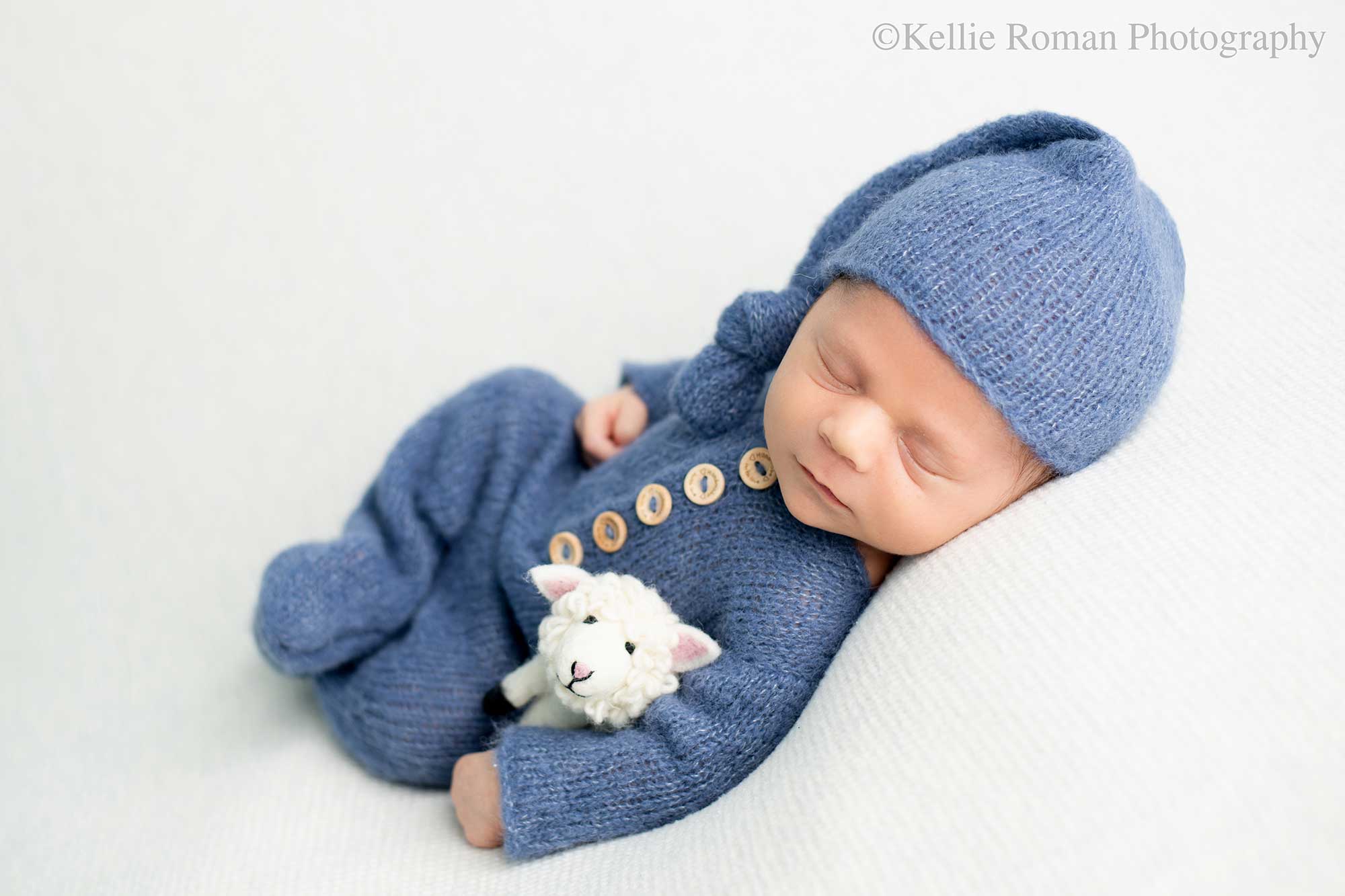 milwaukee newborn photographers. a newborn baby boy is sleeping on his back onto of cream fabric. he has a blue knit footed romper on with wood buttons and a matching hat. he has a felt lamb tucked into his arm.