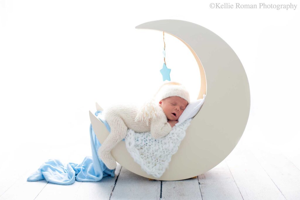milwaukee newborn photographers. a newborn baby boy is sleeping on his belly onto of a cream wood moon prop. there are white and light blue fabrics draped over the moon. the boy has one leg dangling off the blue and the other hand is under his chin. he has his mouth open. the newborn has a cream knit footed romper on with a matching hat. the backdrop is bright white.