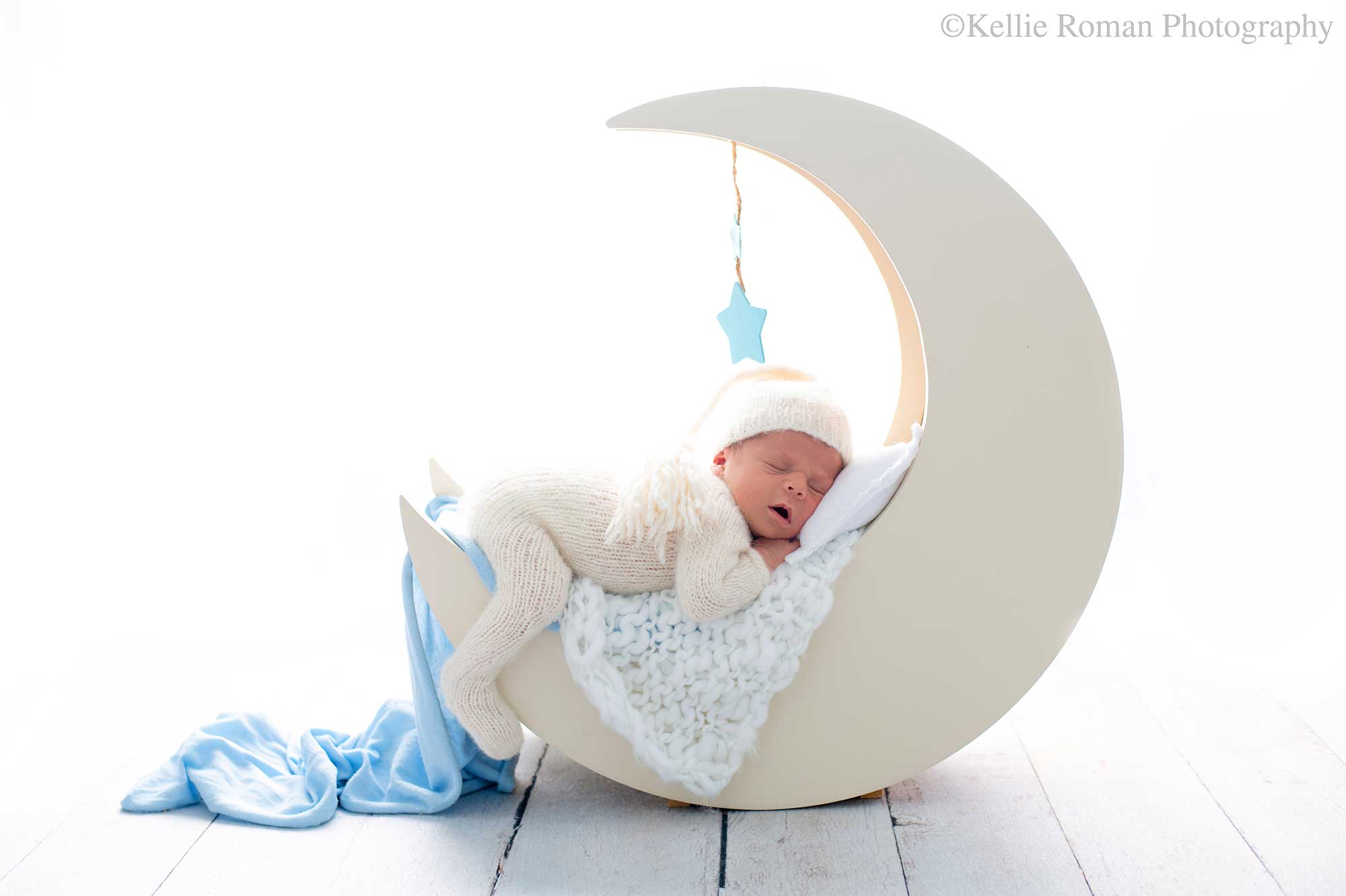 milwaukee newborn photographers. a newborn baby is is asleep on his belly on a cream wood moon. there are fabrics on the moon like a blue swaddle wrap. the newborn is wearing a cream knit footed romper. the backdrop is bright and white.