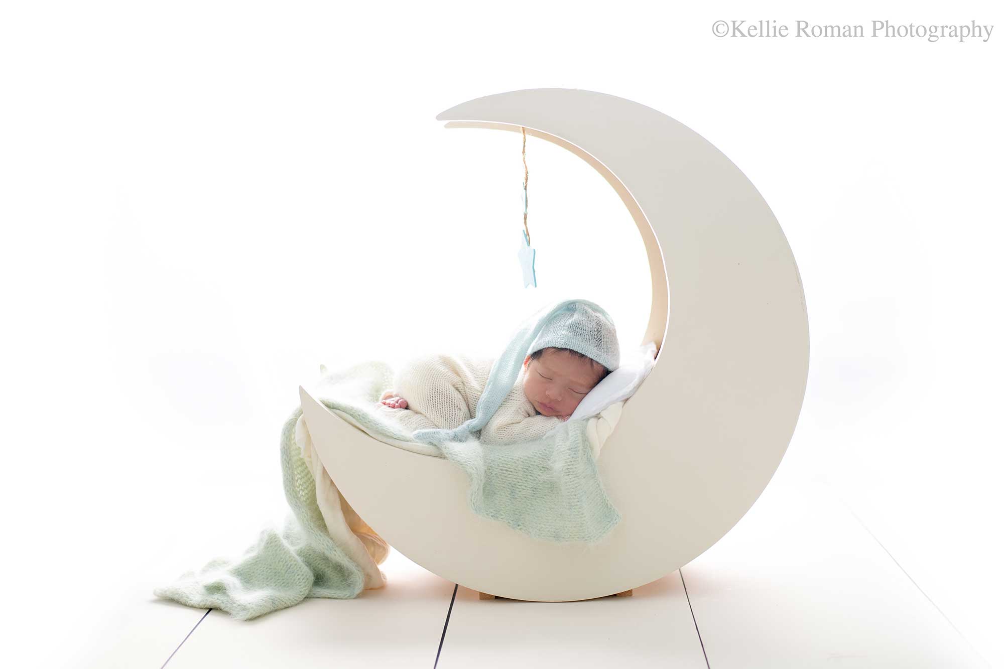newborn photography milwaukee. a newborn boy is asleep on a cream wood moon prop. baby has a cream knit romper on with a blue sleepy hat. there are blue and cream fabric layers on the moon. the backdrop is bright white.