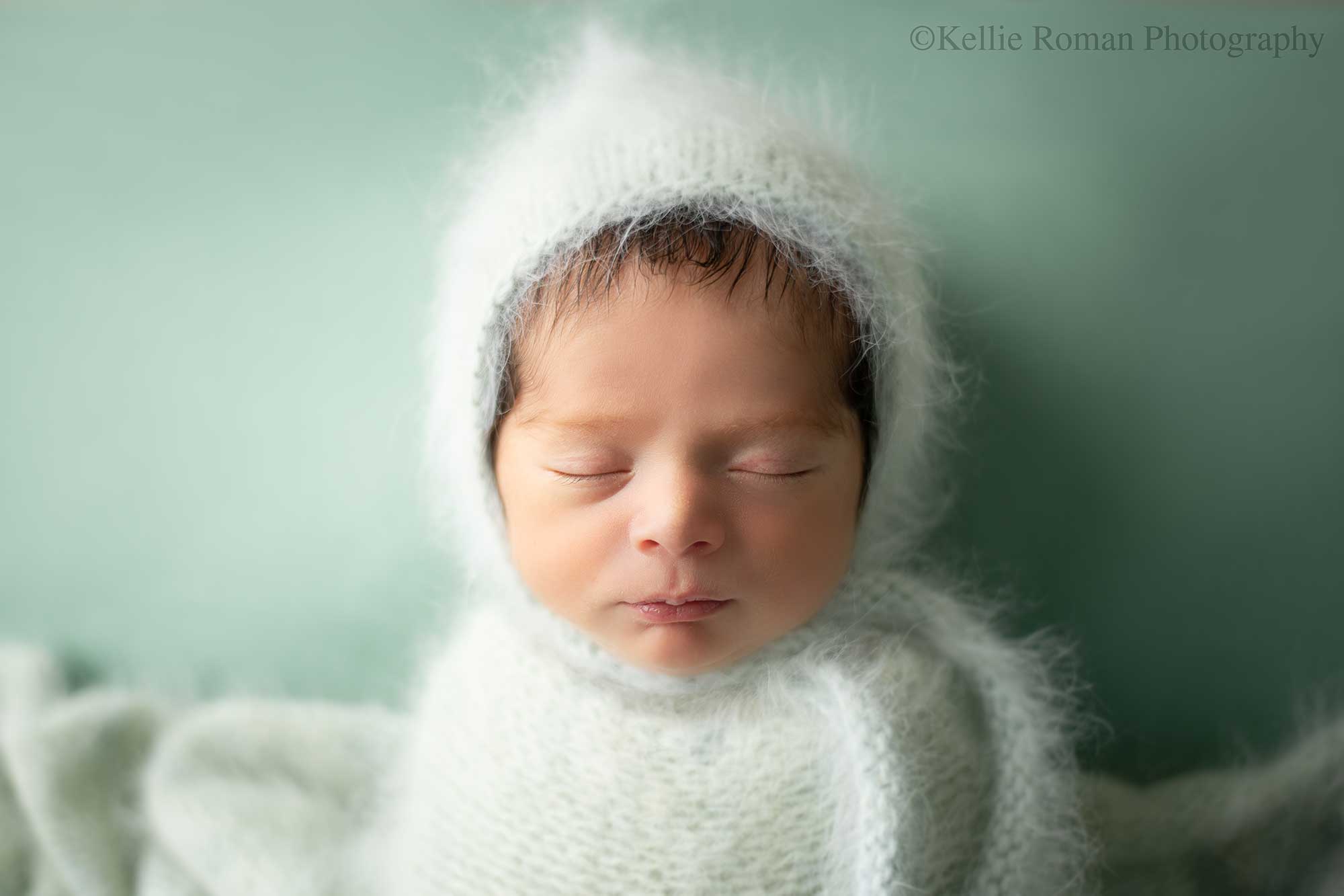 newborn photography milwaukee. a newborn baby boy is asleep on his back on top of teal fabric. he has a fuzzy light teal bonnet on with a matching swaddle over him.