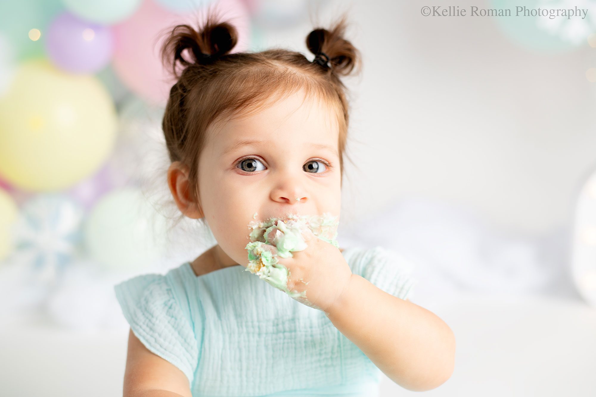Milwaukee pastel cake smash. one year old girl is shoving cake into her mouth. her hand is covered in green frosting. she has a light blue romper on with pig tails. the background in pastel balloon garland. 