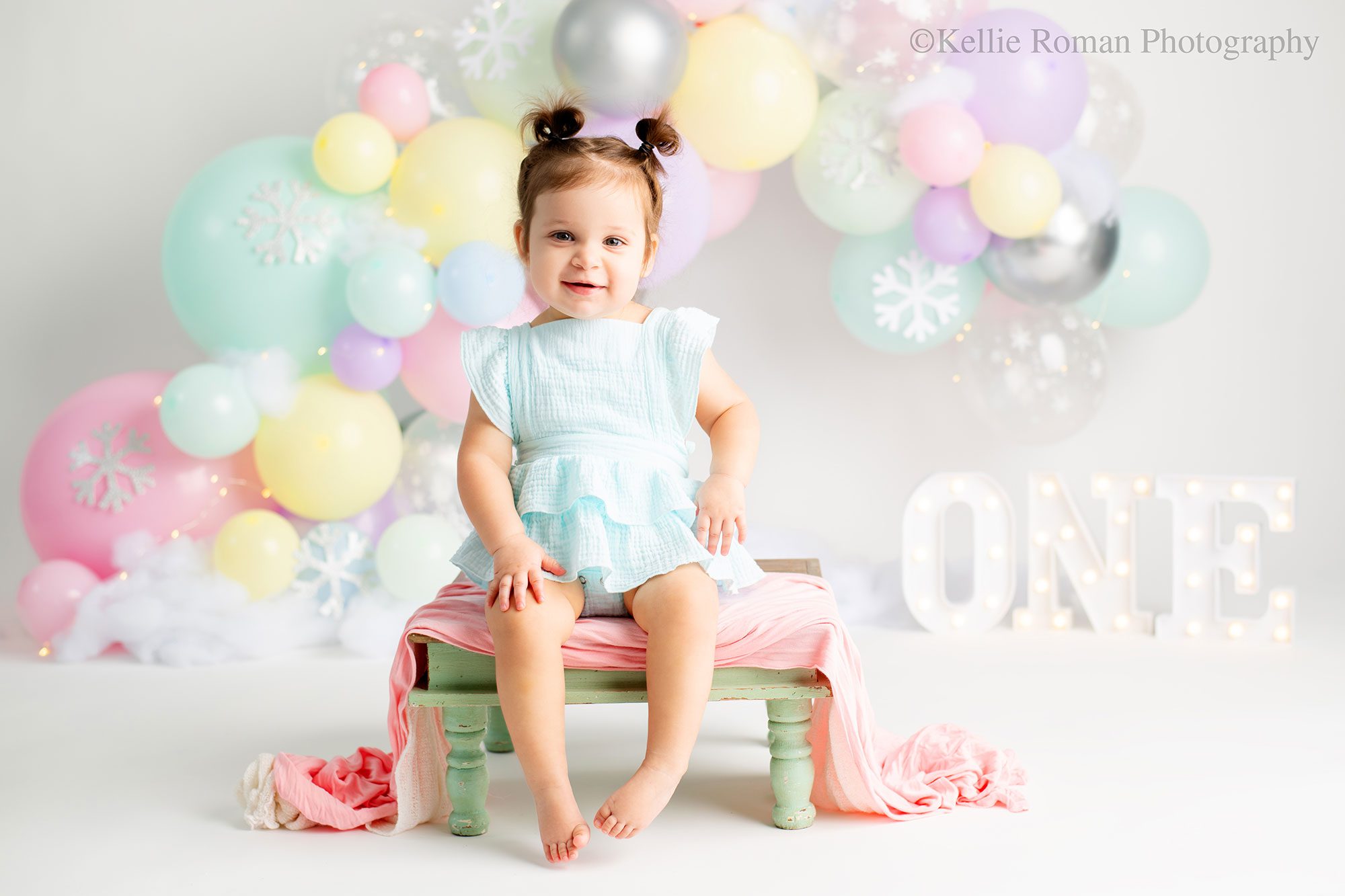 milwaukee pastel cake smash. one year old girl sitting on a teal wood bench with pink fabric over it. she's wearing a light blue romper and has pig tails. she's smiling at the camera. the background is pastel balloon garland with snowflakes and little lights.