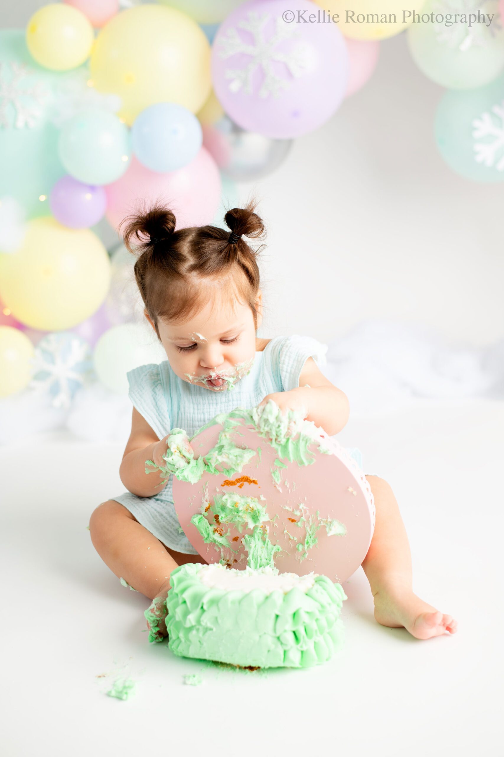 milwaukee pastel cake smash. one year old girl covered in frosting is tipping green cake off a pink cake stand. she has a light blue romper on and pigtails. the background is a custom made pastel balloon garland. 