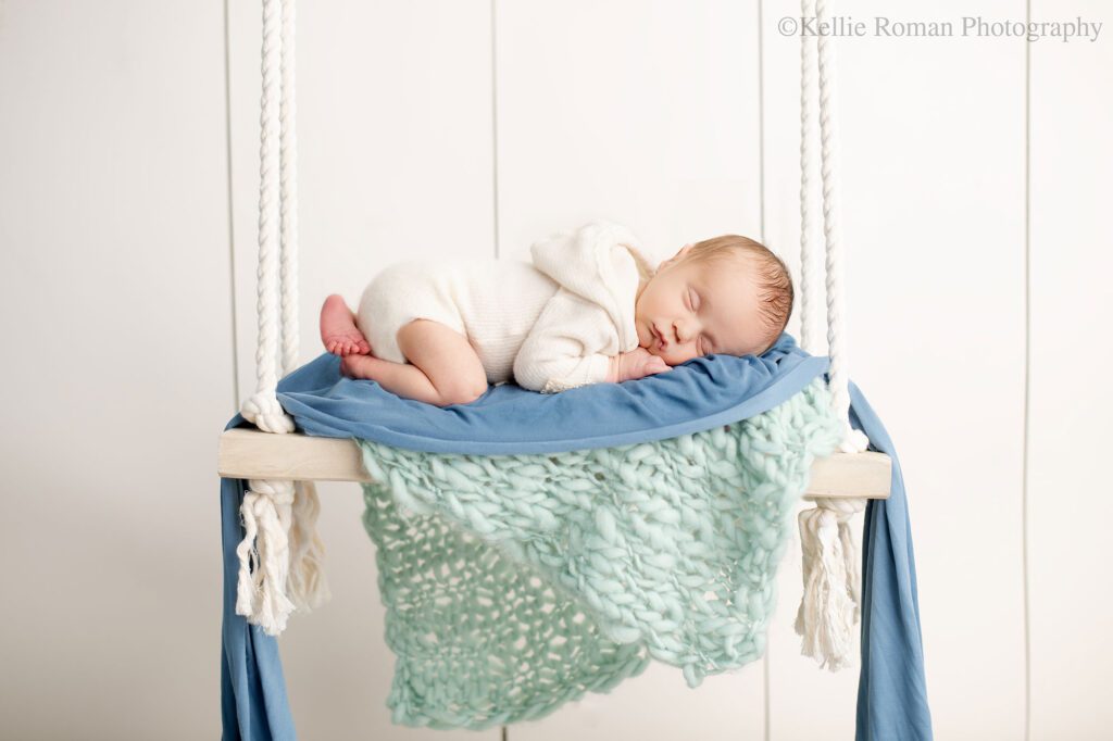 milwaukee newborn pics. newborn baby boy is sleeping on a wood swing that has teal and blue fabric on it. baby has a cream colored romper on.