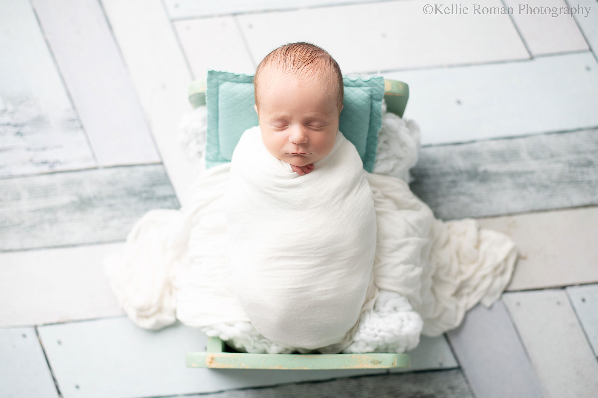 Milwaukee newborn pics. a newborn baby boy is swaddled in a white wrap and is sleeping in a teal wood bed. there is a light blue pillow, and fake blue and cream flooring. 