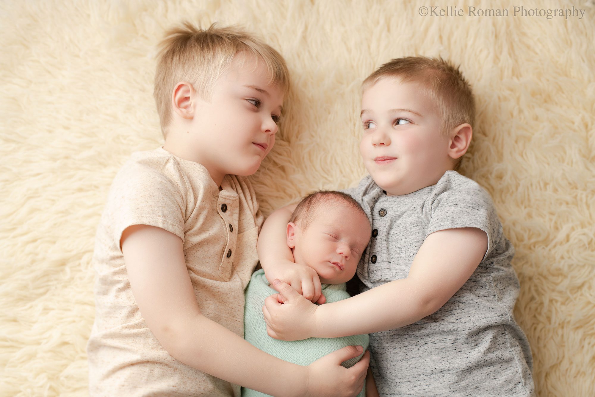 Milwaukee newborn pics. two brothers are looking at each other while laying on a cream fuzzy rug. they are cuddling their newborn brother who's laying in between them on the rug. the baby is asleep. 
