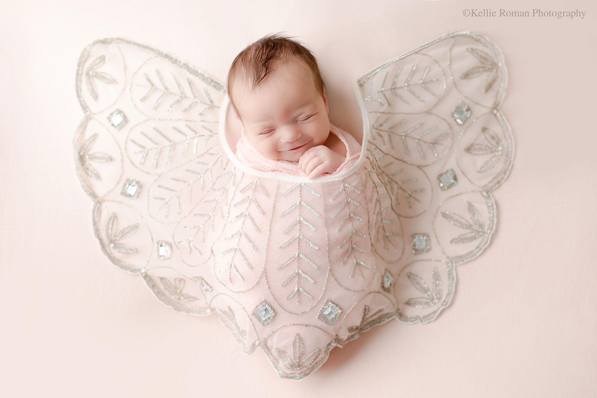 experienced Milwaukee newborn photographer. newborn baby girl is asleep on her back onto of light pink fabric. she's swaddled in a light pink swaddle, with a sheer decorative fabric over her. the fabric is a pattern of silver sequins. the baby is smiling.