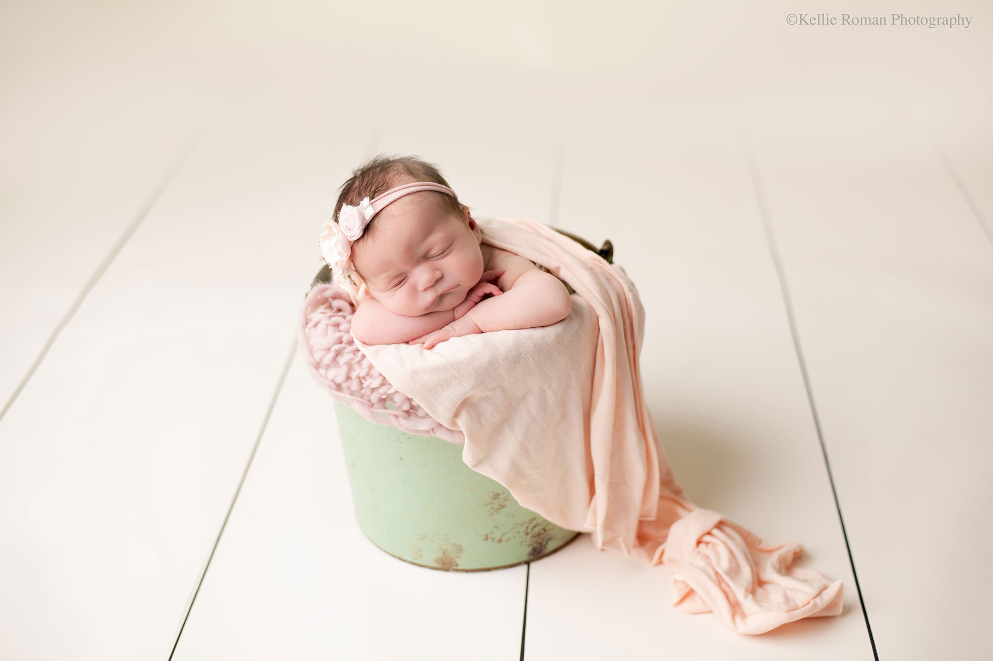 experienced milwaukee newborn photographer. newborn girl is asleep in a bucket with her chin resting on her hands. the bucket is sage green, there are pink fabrics inside. the floor and background are soft cream. 