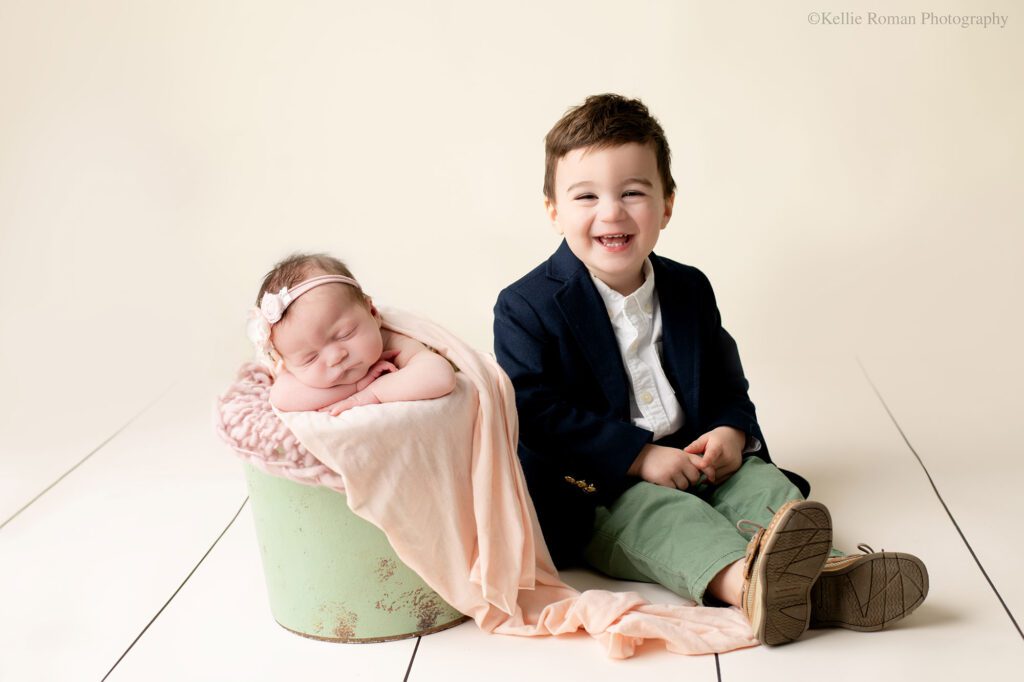 experienced milwaukee newborn photographer. a baby girl is asleep in a sage green bucket, filled with light pink fabrics. her chin is resting on her arms, and she has a floral headband on. her toddler brother is sitting next to her in the bucket and smiling very big. he has sage green pants on with a white shirt, and blue sports coat.