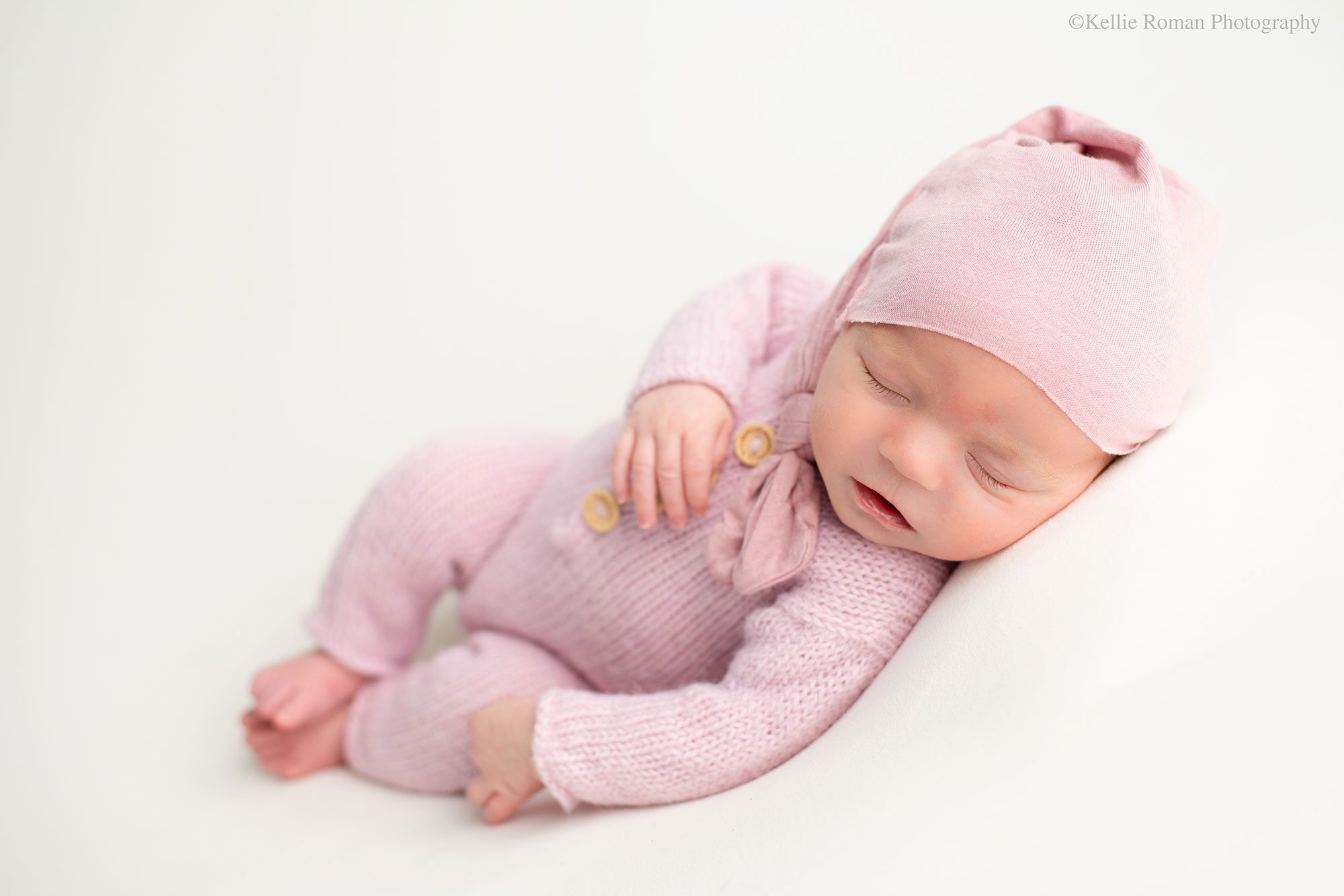 experienced Milwaukee newborn photographer. baby girl is sleeping laying on her side on top of cream fabric. she has a pink knit onside one with wood buttons down the front and a matching pink sleepy cap. 