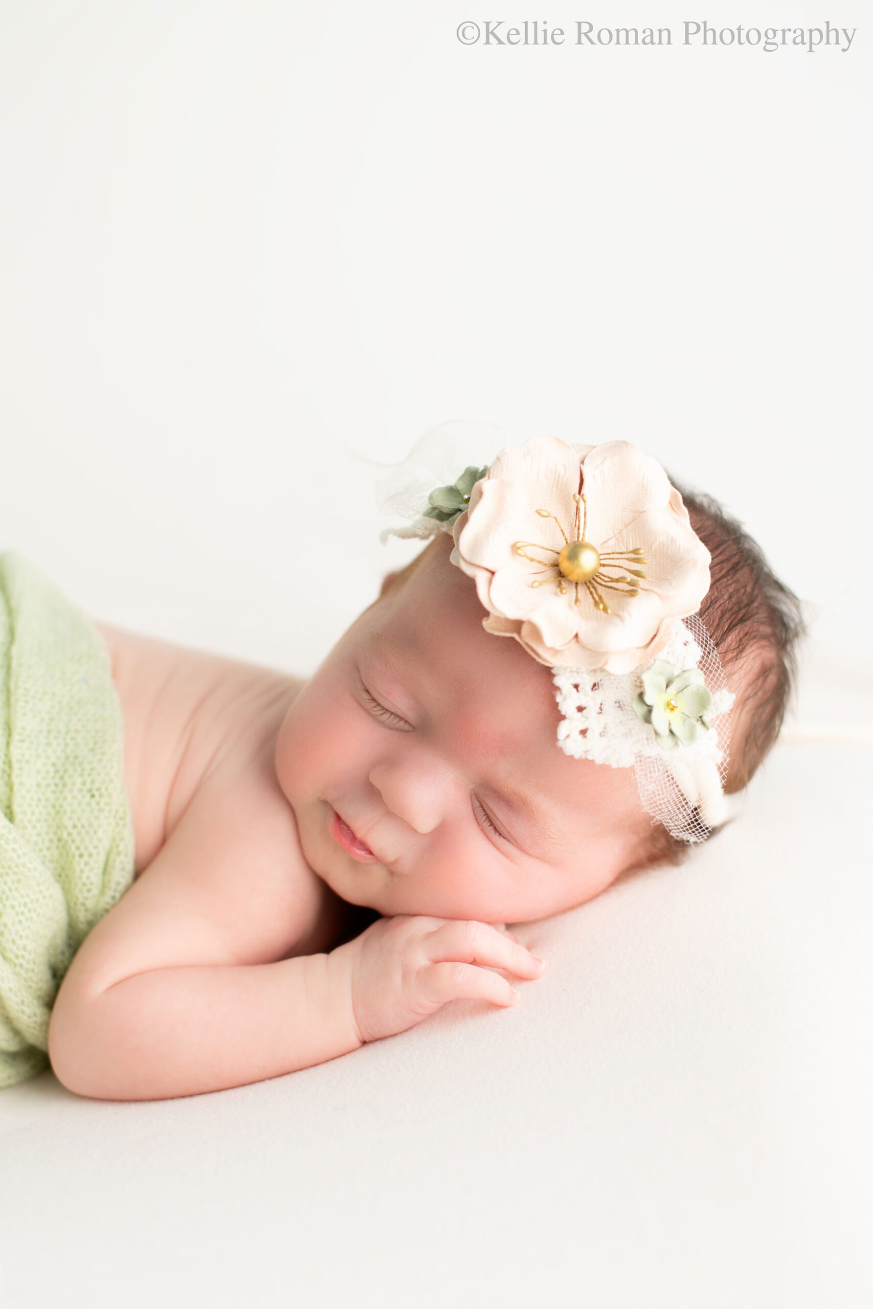 experienced Milwaukee newborn photographer. close up image of a newborn girls face. she's asleep on her belly with her hand resting under her chin. she has a light green fabric over her back and a floral green and cream headband on. 