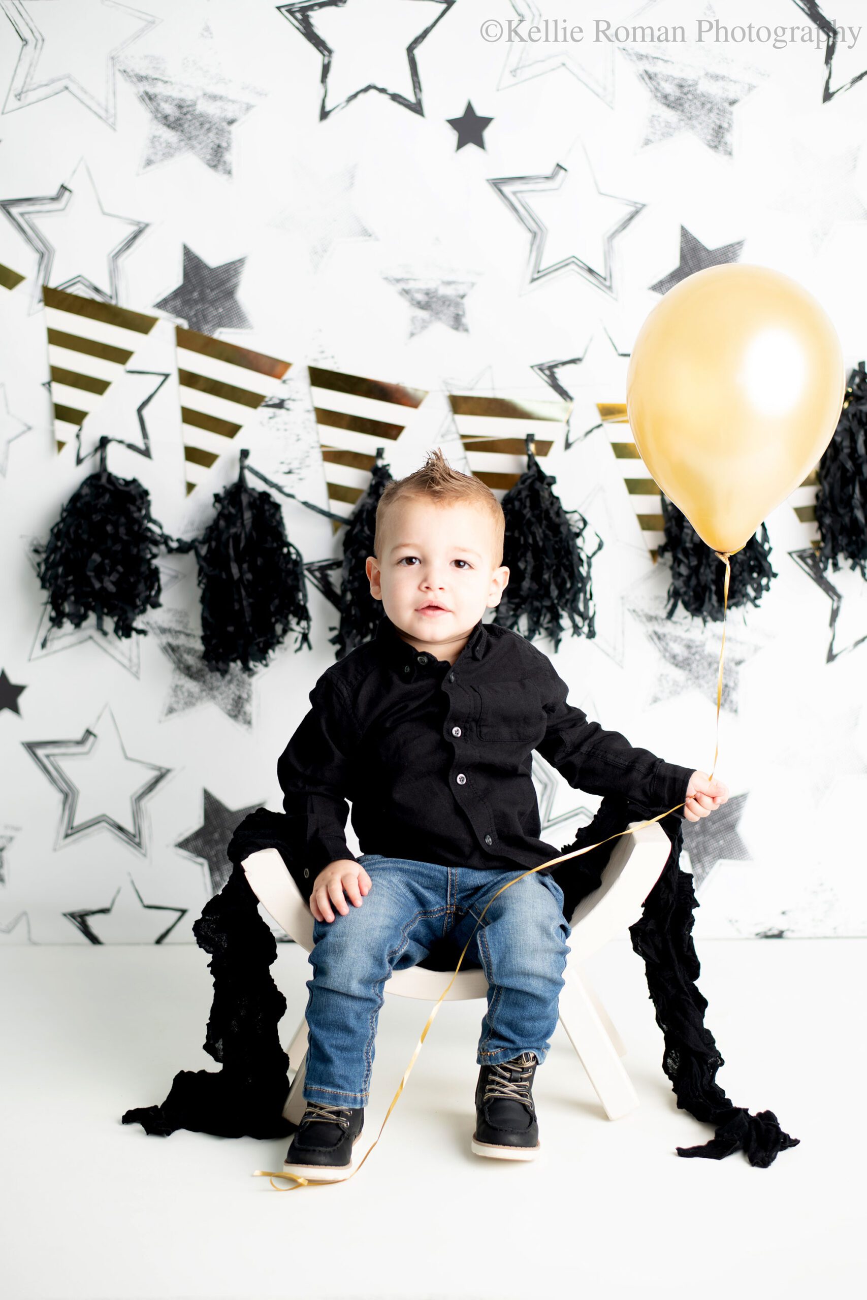milwaukee child photographer. two year old boy in a black shirt with jeans is sitting on white curved bench. he's holding a gold balloon, and looking at the camera. the backdrop is white with black stars, black garland, and gold and white banner. 
