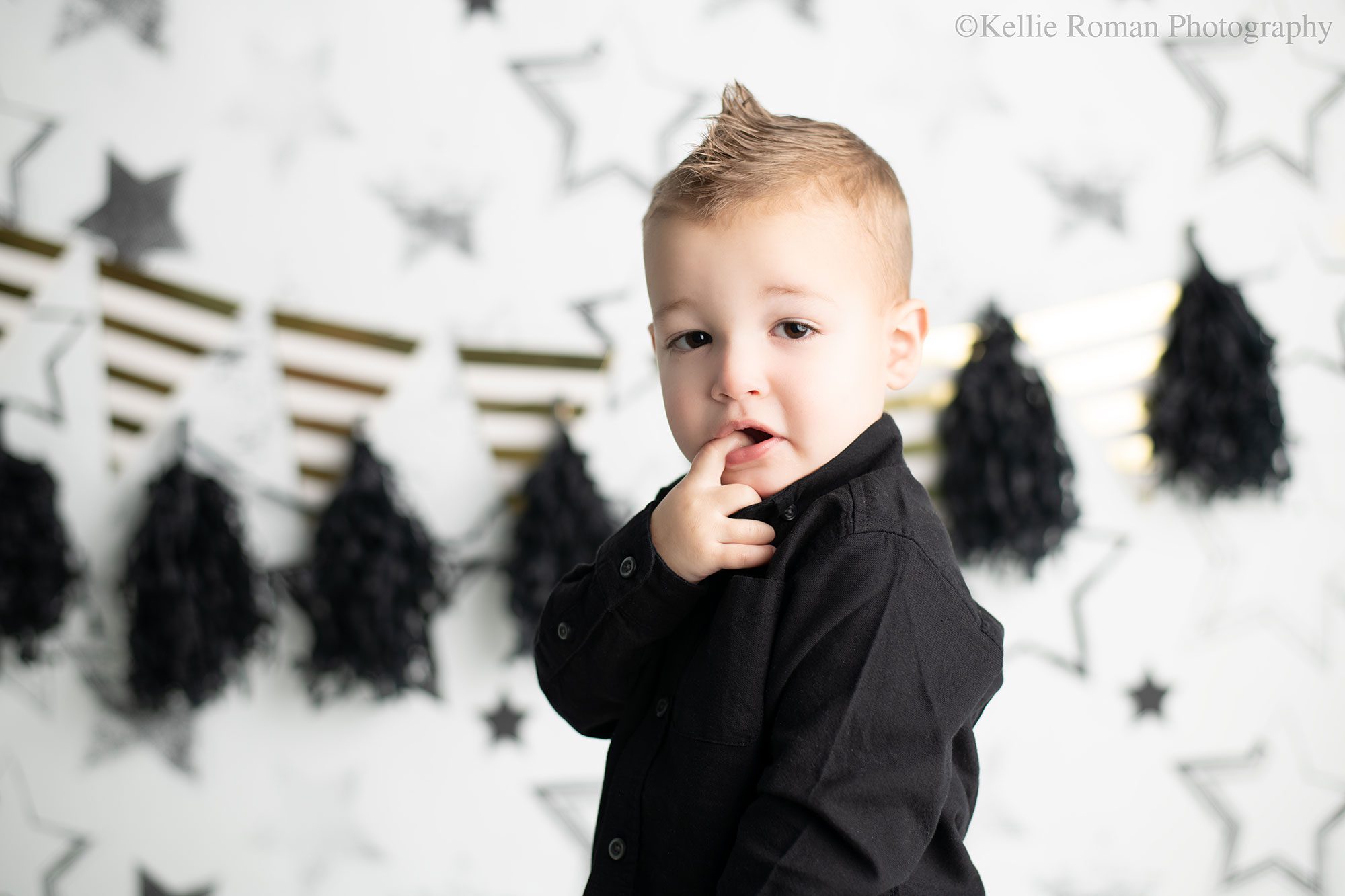 milwaukee child photographer. two year old boy standing with his finger in his mouth looking at camera. he hs a black shirt on and a faux hawk. the backdrop is white with black stars and black and gold banners.