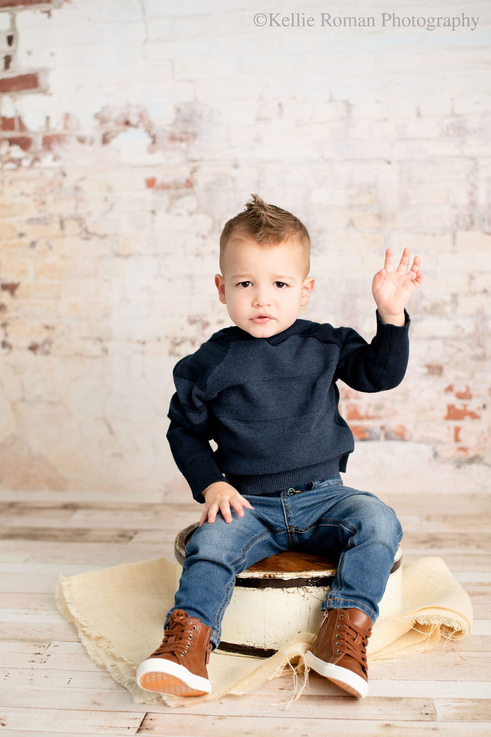 milwaukee child photographer. two-year old boy wearing a navy sweater and jeans with brown boots. he's sitting on a cream, wood bucket infront of a cream, brick backdrop. he's raising one hand and looking concerned at the camera. 