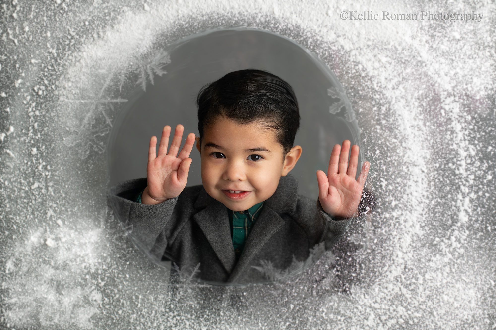 birthday photographer milwaukee. young boy is standing behind glass with snow all over it holding his hands up. he's smiling big, and wearing a grey coat and has black hair.