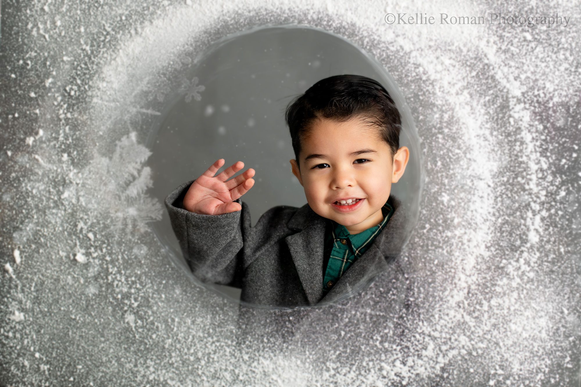 birthday photographer milwaukee. a young boy is standing behind piece of glass with snow sprayed on it waving. he is smiling with dark black hair and a grey jacket. 