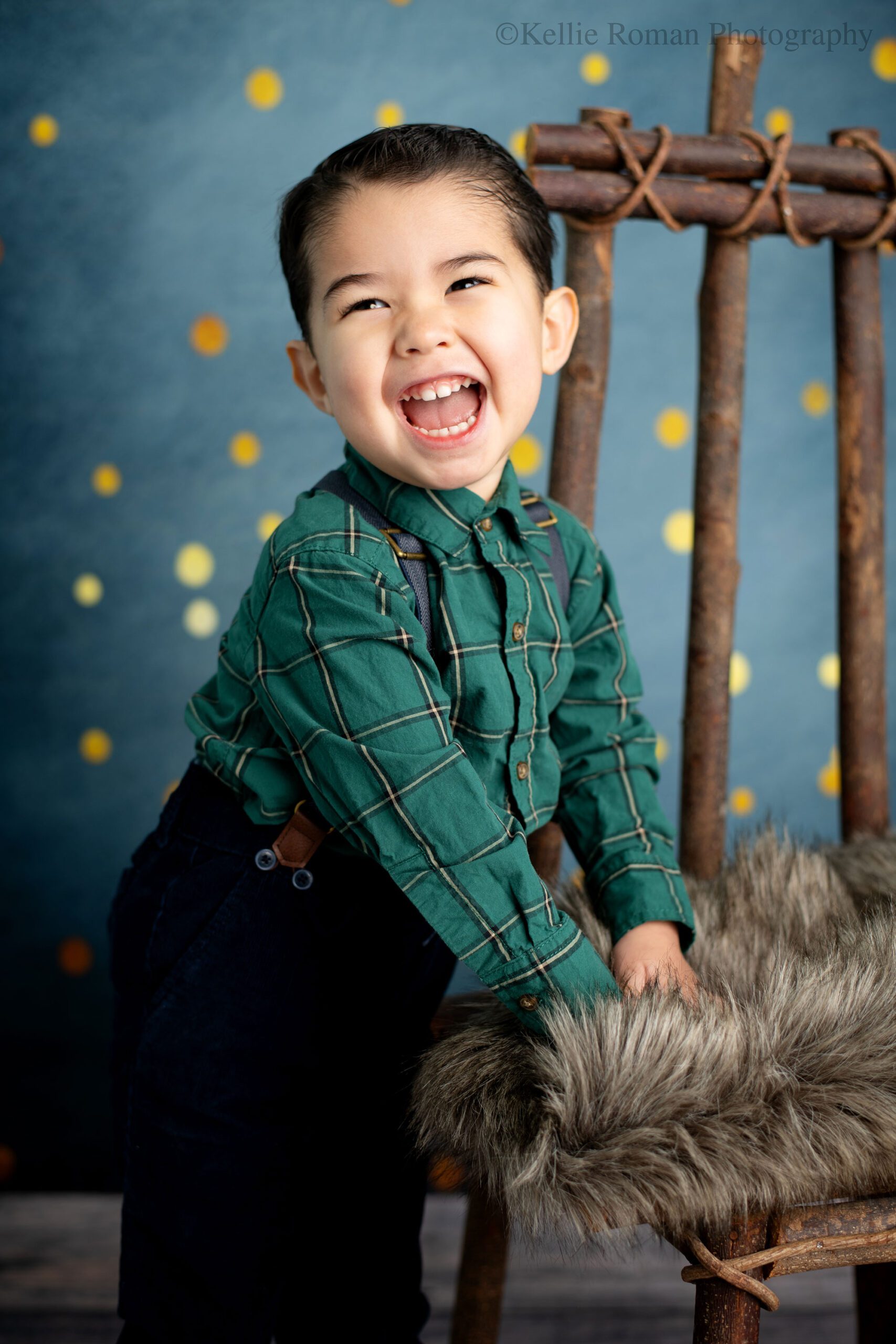 birthday photographer milwaukee. a young boy is looking to the side and smiling really big. he has a green flannel shirt on with navy suspenders and corduroy pants. he's leaning on a wood chair with brown fur over it. the backdrop is navy navy with gold polka dots. 
