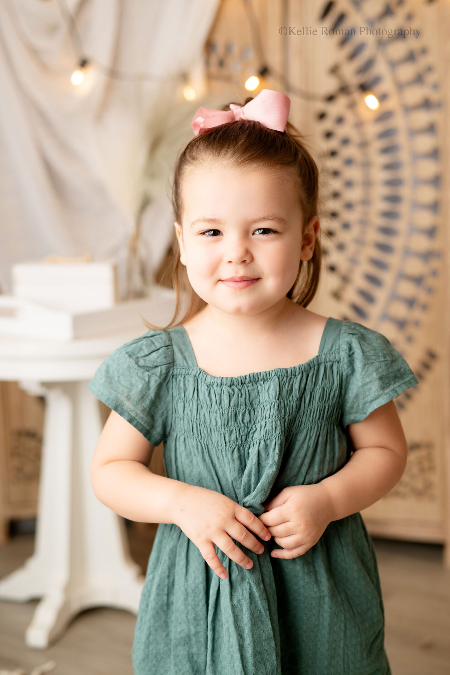 photographer near me. three year old girl is looking at camera and smiling slightly. she has an olive green dress on and is holding onto it a little with her hands. she has a light pink bow in her hair. the background is wood decorative piece and a white wood table.