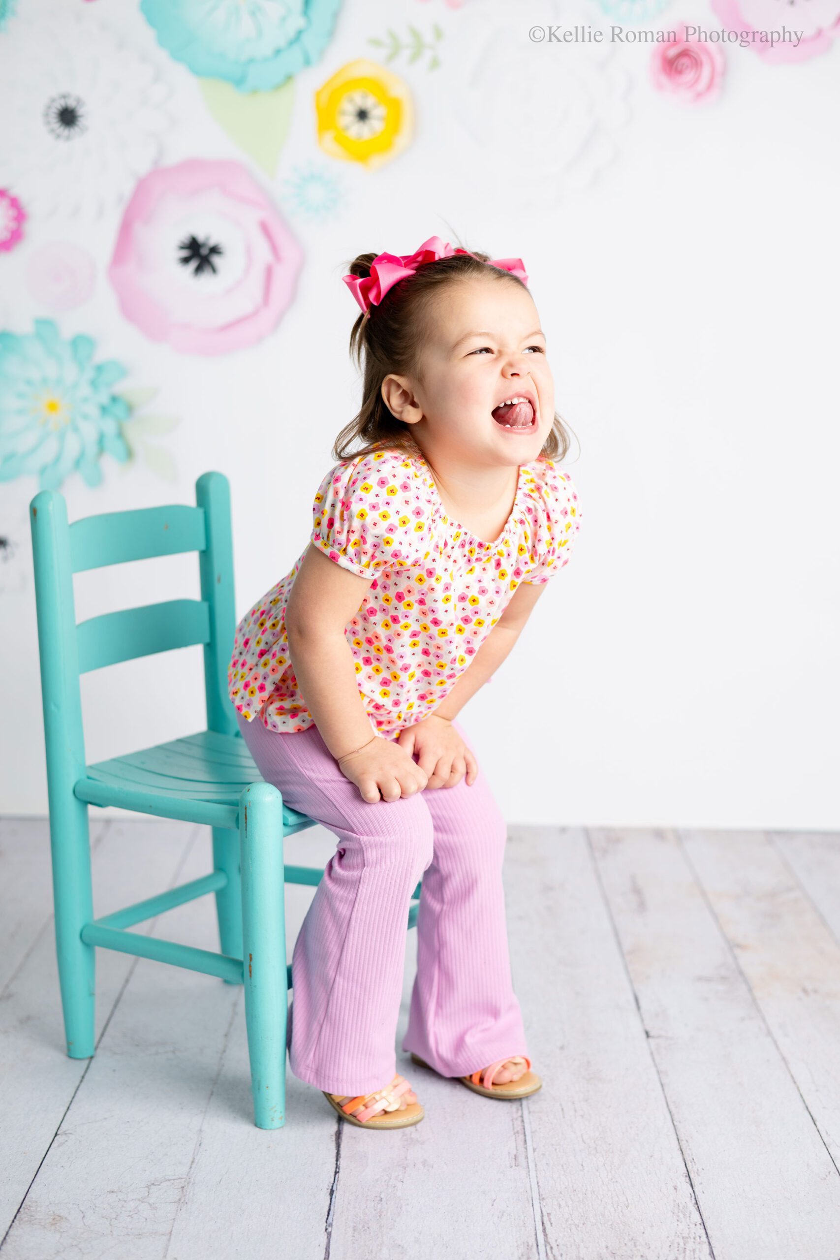 photographer near me. three year old girl is looking off to the side making a silly face with her tongue sticking out. she has a flower shirt on with light purple pants, and pink bows in her pigtails. she's half sitting on a teal chair. the background is white with pink yellow and teal flowers. 