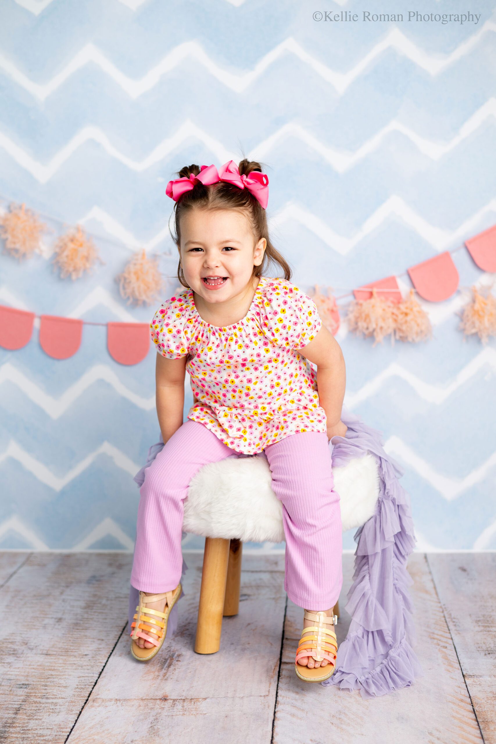 photographer near me. three year old girl smiling big sitting on a white fluffy stool. she has on a colorful shirt with flowers, light purple pants, pink bows in her pig tails and sandals. the backdrop is light blue zigzag with pink and peach banners. 