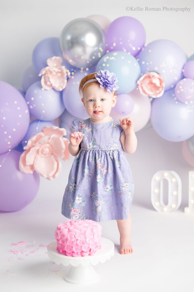 custom cake smash photography milwaukee. cake smash photographers near me. greendale, oak creek, Franklin, Waukesha, racine, Ozaukee cake smash photographer. one year old girl wearing purple dress is smashing pink cake. the background is a balloon garland of purple pink and silver balloons. 